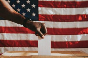 A black hand dropping a ballot in a voting box. An American flag hangs in the background.