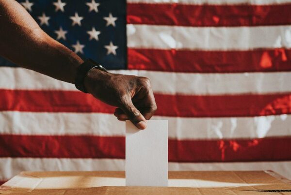 A black hand dropping a ballot in a voting box. An American flag hangs in the background.