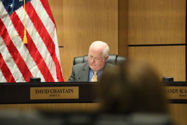 A sad man slumped over in his chair ignoring a public commenter. There is a name plate in front of him that says David Chastain
