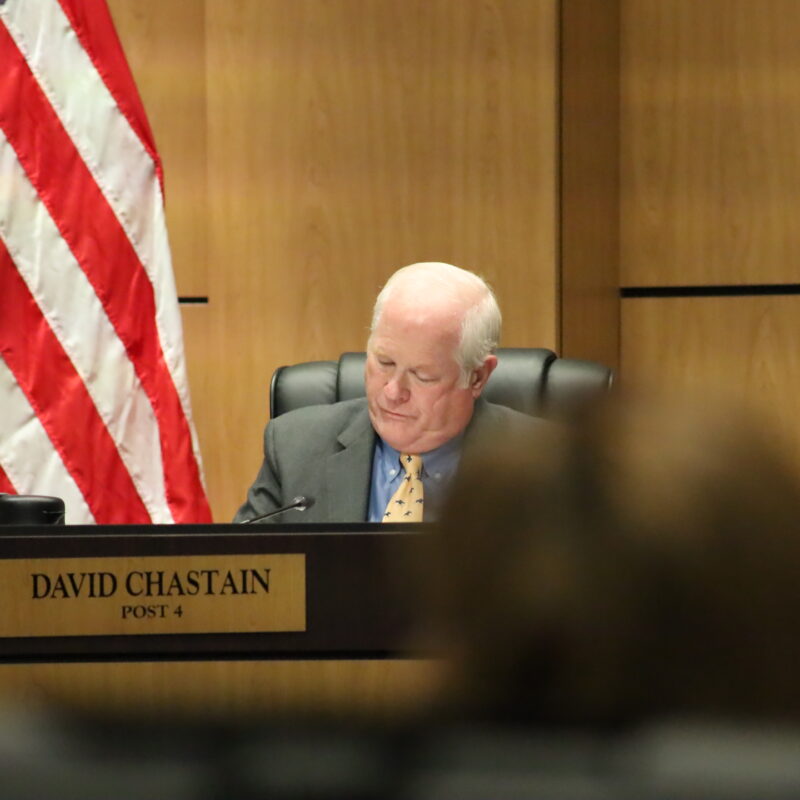 A sad man slumped over in his chair ignoring a public commenter. There is a name plate in front of him that says David Chastain